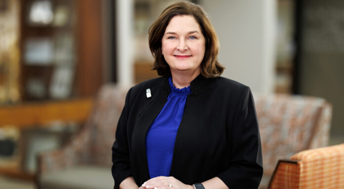 A woman in a blue blouse and black blazer stands indoors, smiling at the camera with hands folded.
