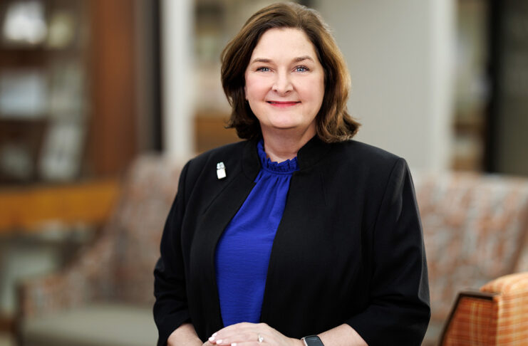 A woman in a blue blouse and black blazer stands indoors, smiling at the camera with hands folded.
