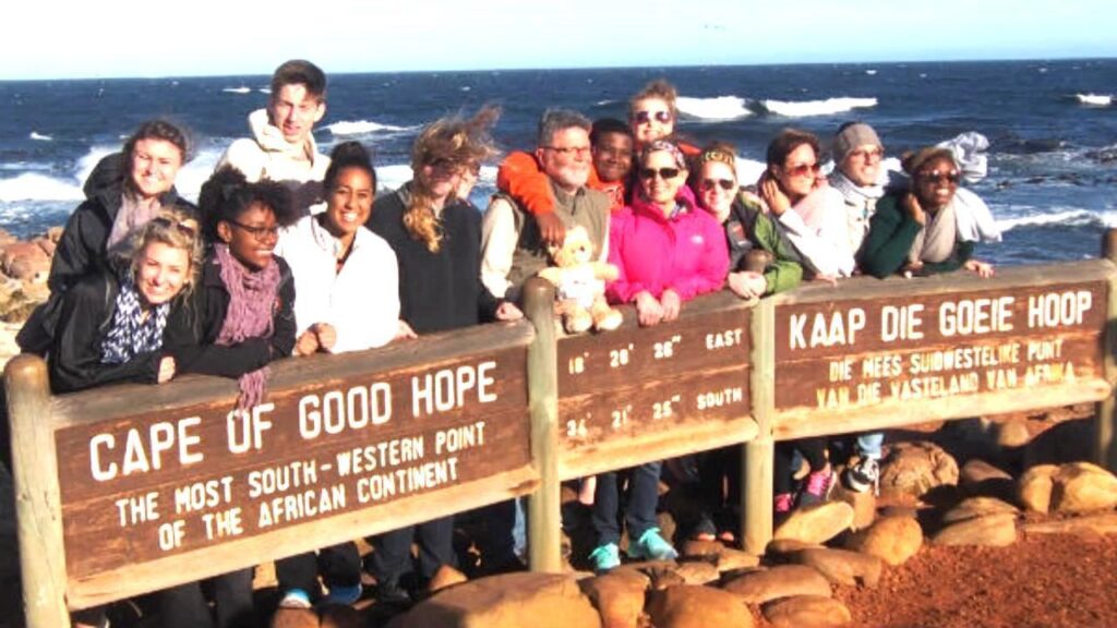 A group of people pose behind signs at the Cape of Good Hope, with the ocean and rocky shore in the background.
