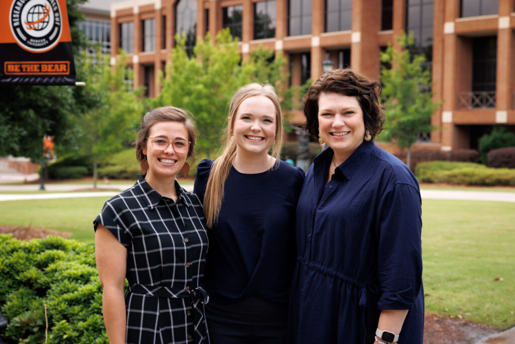 Three women stand together smiling outdoors in front of a university building and a Be The Bear banner.