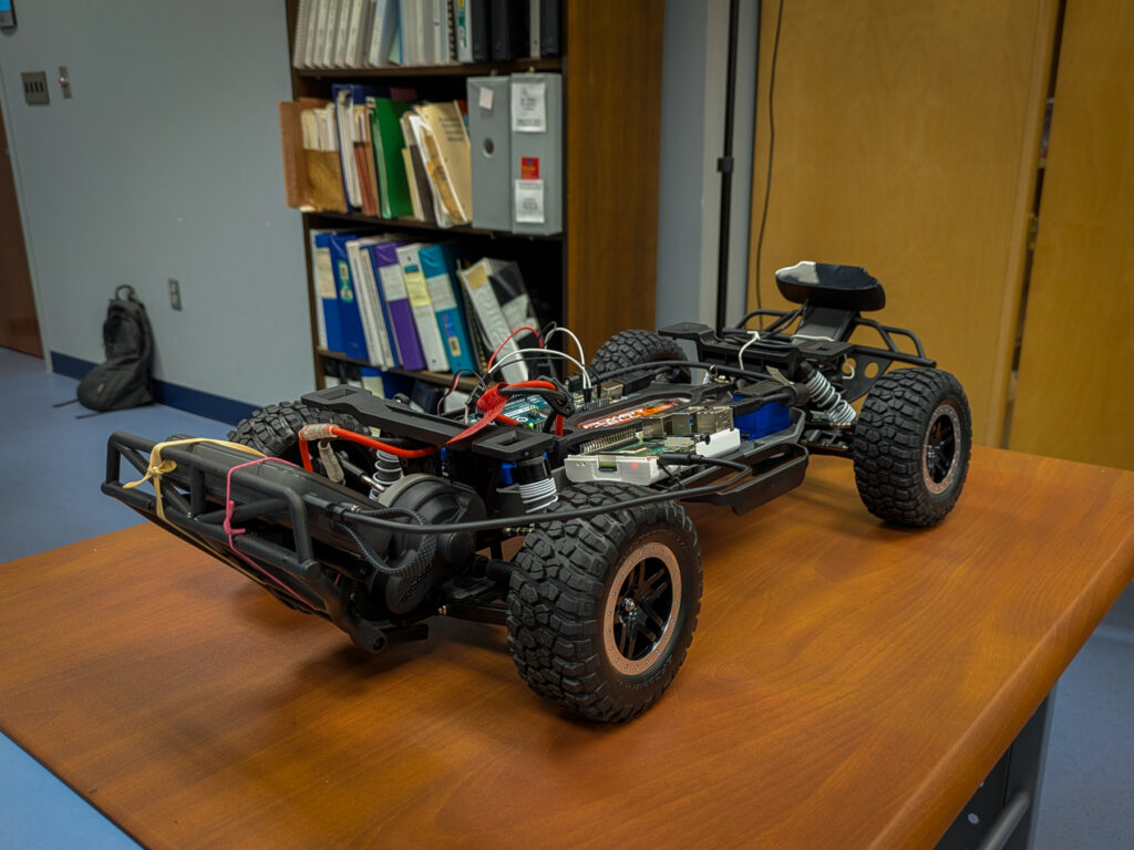 A partially assembled remote-control car with exposed electronics sits on a wooden desk in an office setting.