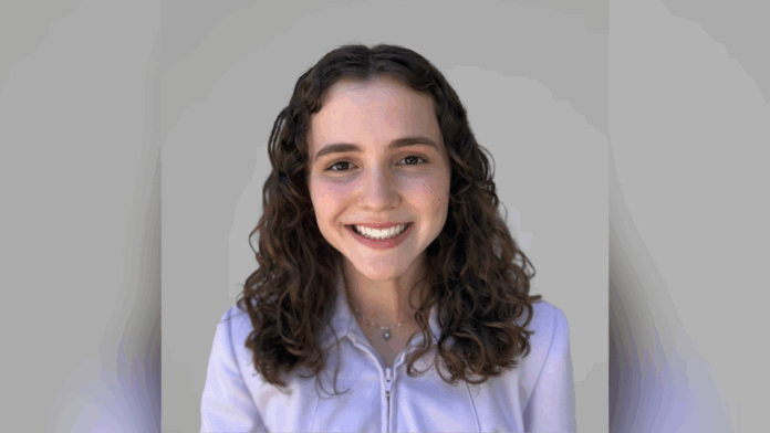 Young woman with curly brown hair smiles at the camera, wearing a light purple zip-up jacket against a plain gray background.