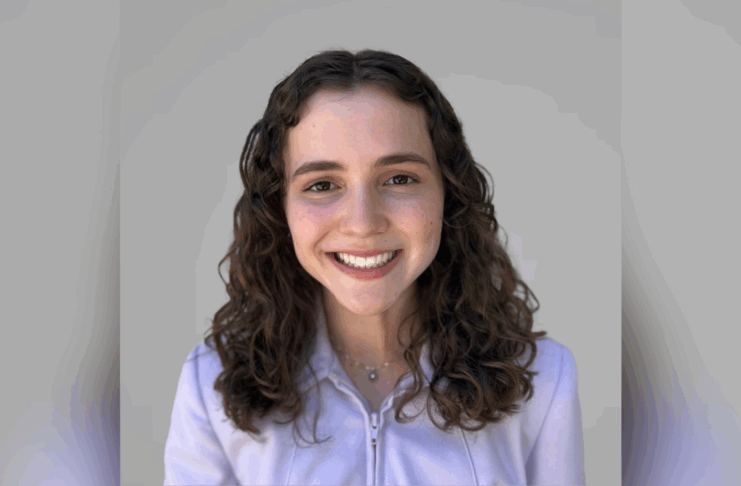 Young woman with curly brown hair smiles at the camera, wearing a light purple zip-up jacket against a plain gray background.