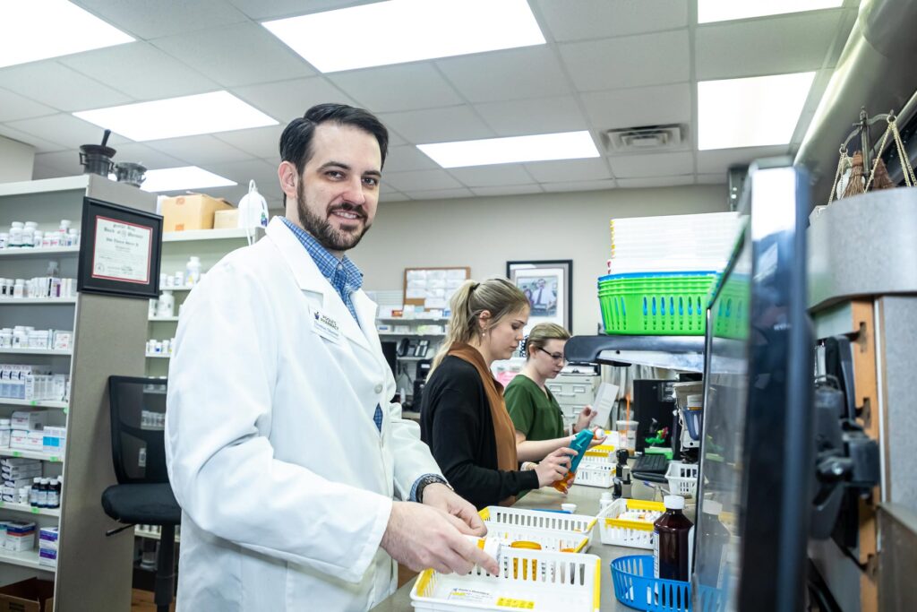A pharmacist in a white coat works at a counter with two colleagues organizing medications in a pharmacy.