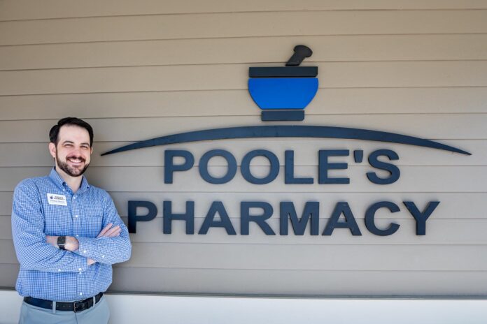 Dr. Thomas Sherrer stands smiling with arms crossed next to a sign reading Poole's Pharmacy on an exterior wall.