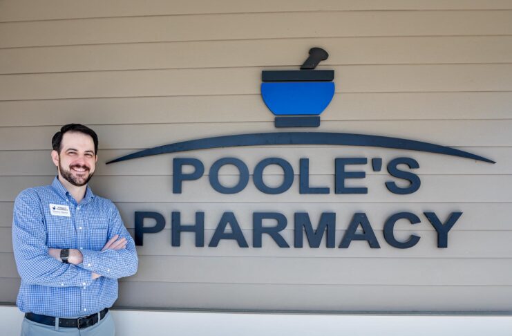 Dr. Thomas Sherrer stands smiling with arms crossed next to a sign reading Poole's Pharmacy on an exterior wall.