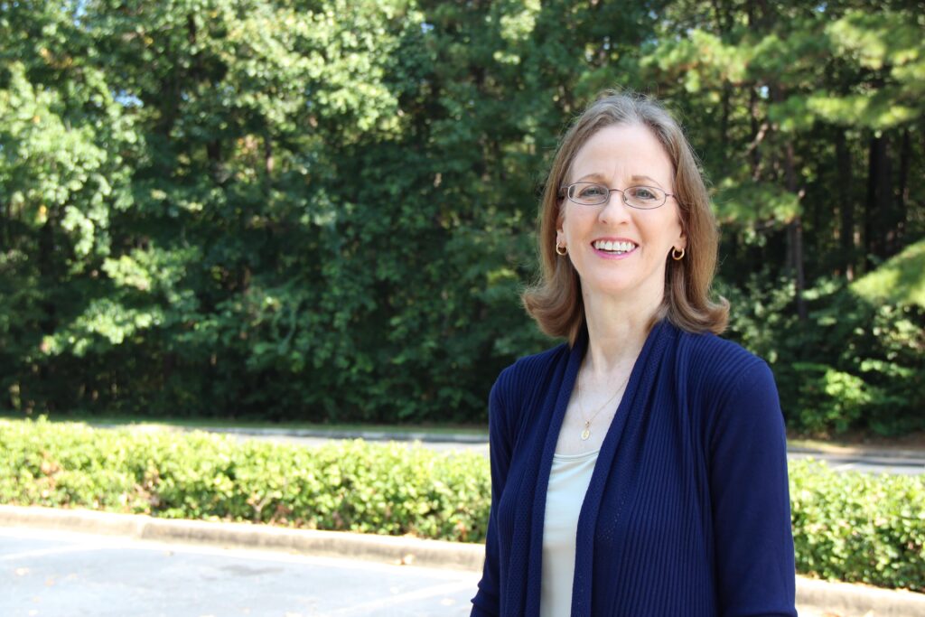 A woman with glasses stands outdoors in a parking lot, smiling, with trees and greenery in the background.