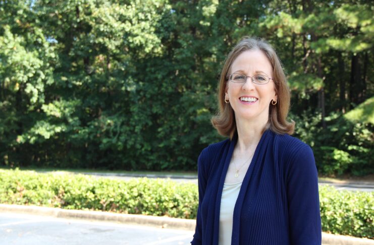 A woman with glasses stands outdoors in a parking lot, smiling, with trees and greenery in the background.