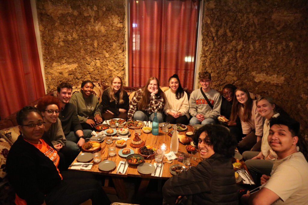 A group of people sit around a low table, sharing a meal in a warmly lit restaurant with earthen walls.