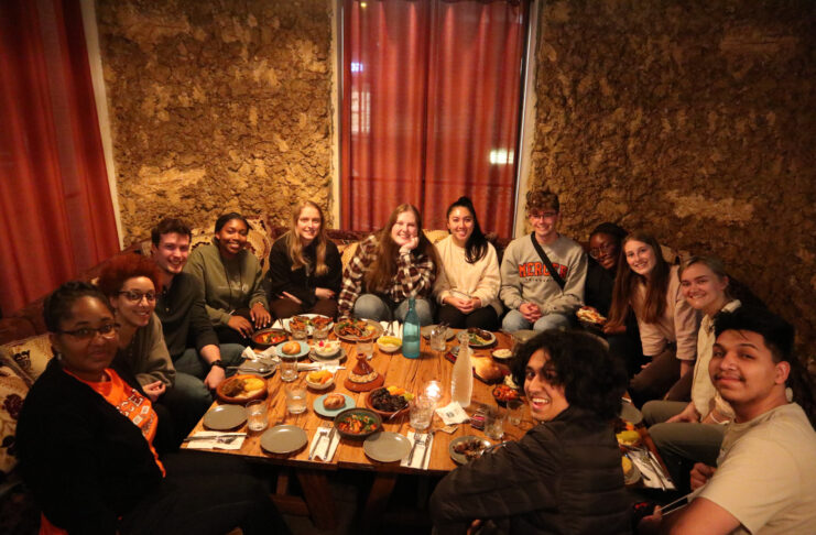A group of people sit around a low table, sharing a meal in a warmly lit restaurant with earthen walls.
