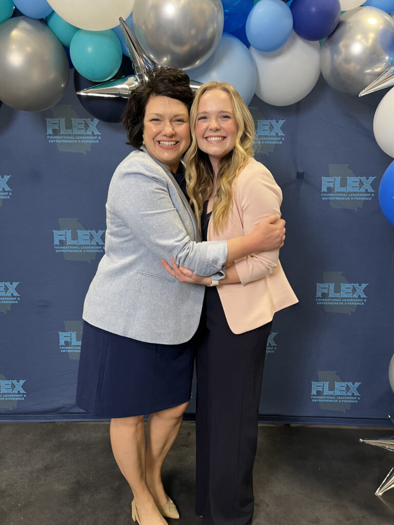 Two women stand smiling and hugging in front of a blue FLEX event backdrop with balloons.