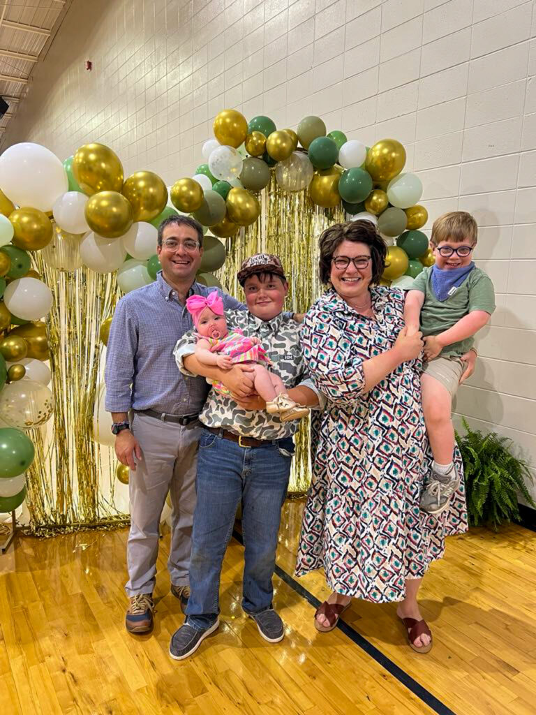 Two adults and three children pose together in front of a gold and white balloon arch in a gymnasium.