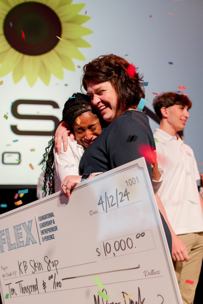 Two women embrace while holding a large $10,000 check on stage as confetti falls.