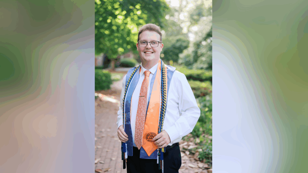 A young person in a white shirt and tie stands outdoors wearing graduation cords and a medal, smiling at the camera.