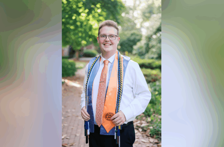 A young person in a white shirt and tie stands outdoors wearing graduation cords and a medal, smiling at the camera.