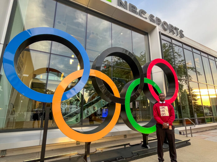 n'kyzie-hawkins Person stands beside large Olympic rings sculpture outside an NBC Sports building.