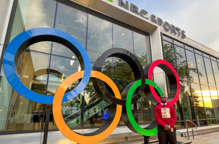 Person stands beside large Olympic rings sculpture outside an NBC Sports building.