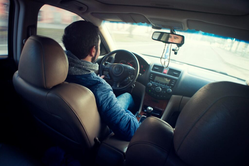 A man in a blue jacket drives a car, viewed from the back seat, with his hands on the steering wheel.