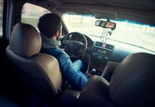 The most important people this summer may be those who help you get home | Dr. Greg DeLoach A man in a blue jacket drives a car, viewed from the back seat, with his hands on the steering wheel.