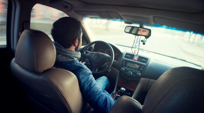 The most important people this summer may be those who help you get home | Dr. Greg DeLoach A man in a blue jacket drives a car, viewed from the back seat, with his hands on the steering wheel.