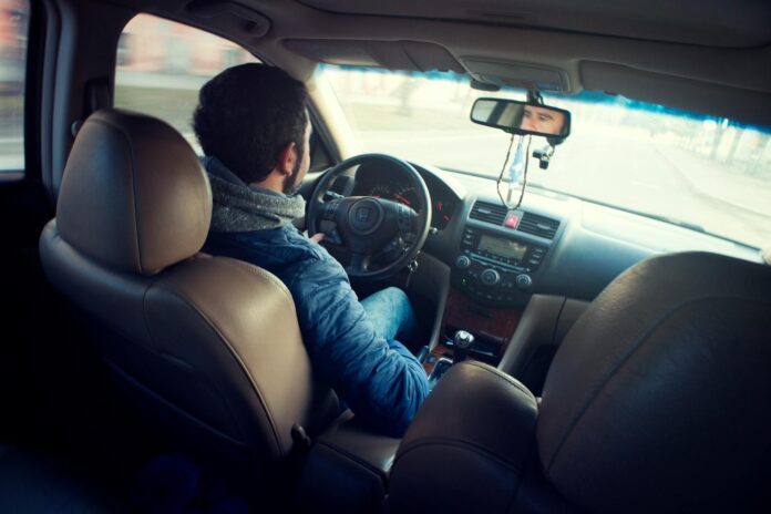 A man in a blue jacket drives a car, viewed from the back seat, with his hands on the steering wheel.