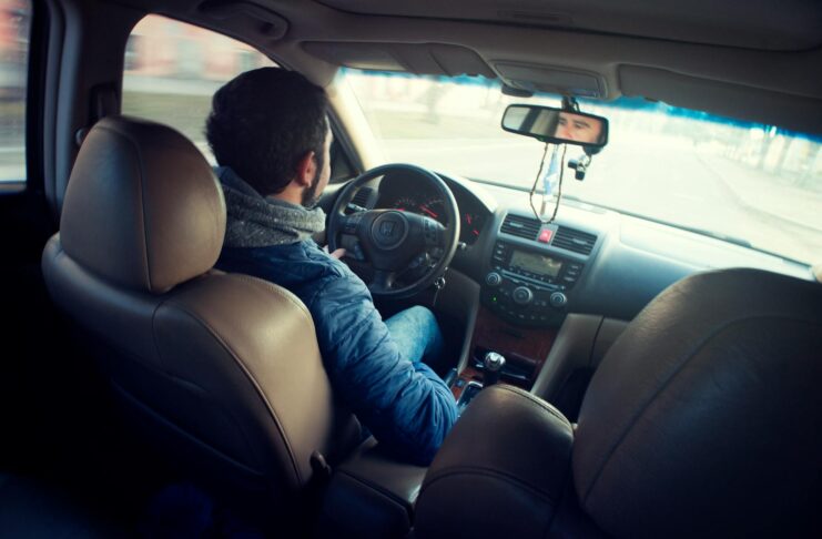 A man in a blue jacket drives a car, viewed from the back seat, with his hands on the steering wheel.