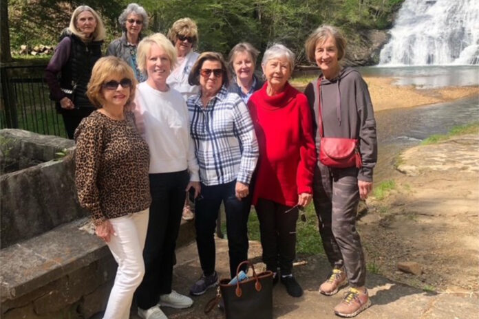 Group of women posing outdoors near a waterfall and a rocky path, with trees and water in the background.