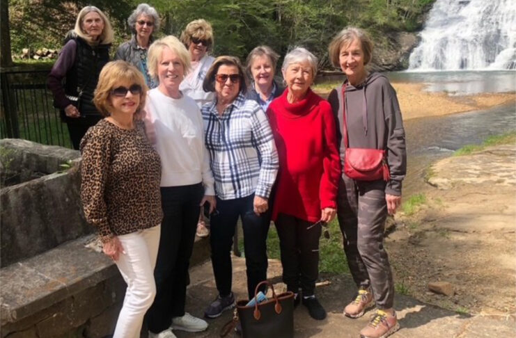 Group of women posing outdoors near a waterfall and a rocky path, with trees and water in the background.