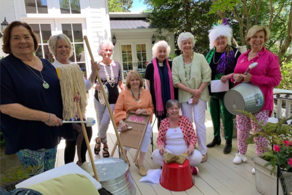 A group of women pose together on a sunlit deck, many holding household items or containers as props.