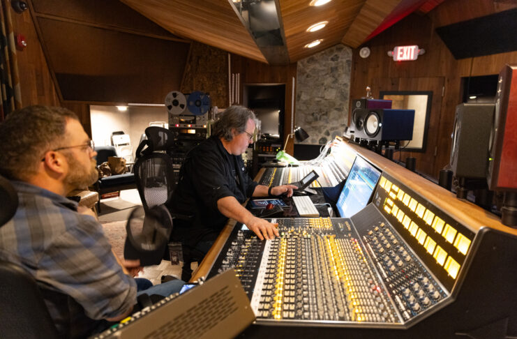 Two men work at a large audio mixing console in a recording studio with wood-paneled walls and various audio equipment.