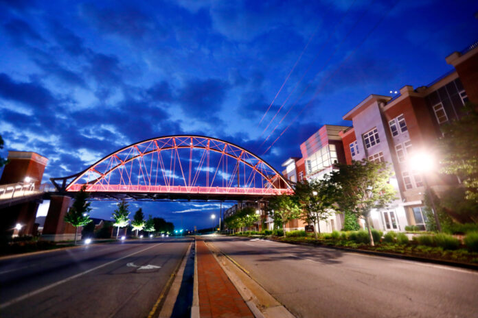 A lit pedestrian bridge spans across an empty street between modern buildings at dusk under a cloudy blue sky.