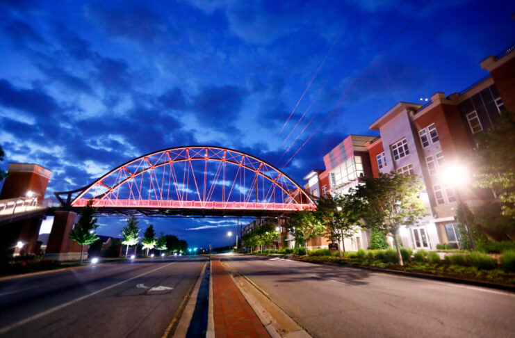 A lit pedestrian bridge spans across an empty street between modern buildings at dusk under a cloudy blue sky.