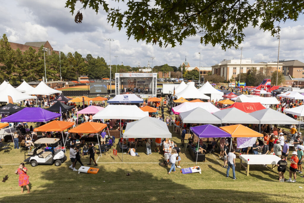 Crowd gathers under colorful tents at an outdoor festival on a grassy field with a stage in the background.