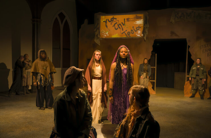 Actors in a dramatic stage scene stand under warm lighting, with a woman in purple at the center and others in muted costumes.