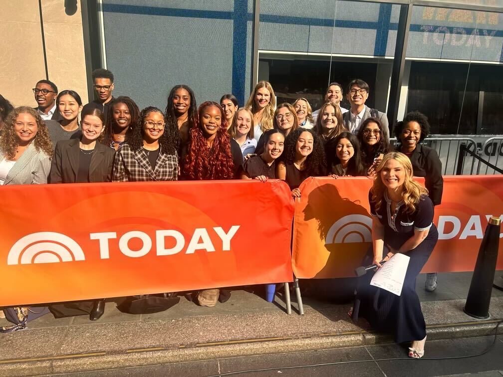 A group of people posing and smiling behind a TODAY show banner outdoors, with a host kneeling in front.