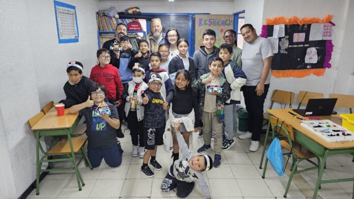 MOM colombia 3 A group of children and adults pose and smile in a classroom filled with desks, colorful posters, and bookshelves.