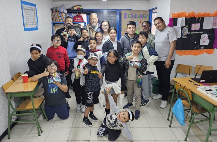 A group of children and adults pose and smile in a classroom filled with desks, colorful posters, and bookshelves.