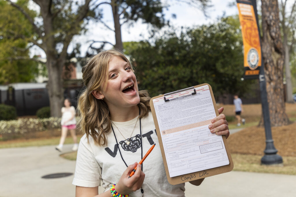 A person outdoors holds a clipboard with a form, smiling and pointing at it with an orange pen.
