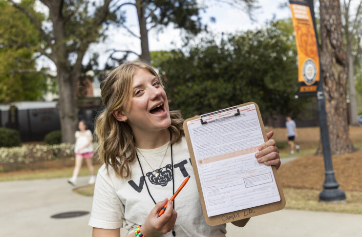 A person outdoors holds a clipboard with a form, smiling and pointing at it with an orange pen.