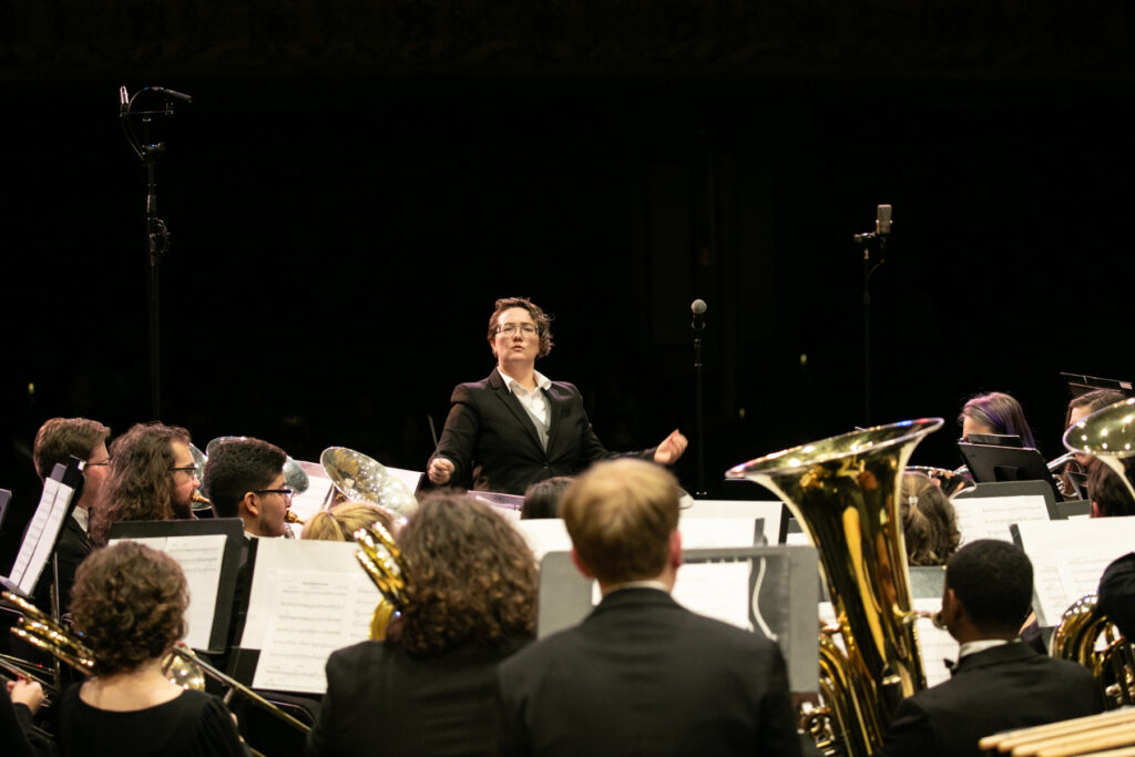 A conductor leads a seated brass and woodwind ensemble during a live concert performance on stage.