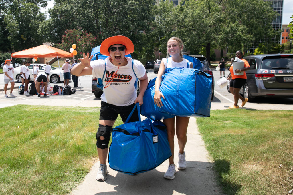 Two students carrying large blue bags walk on a campus sidewalk during a sunny move-in day event.