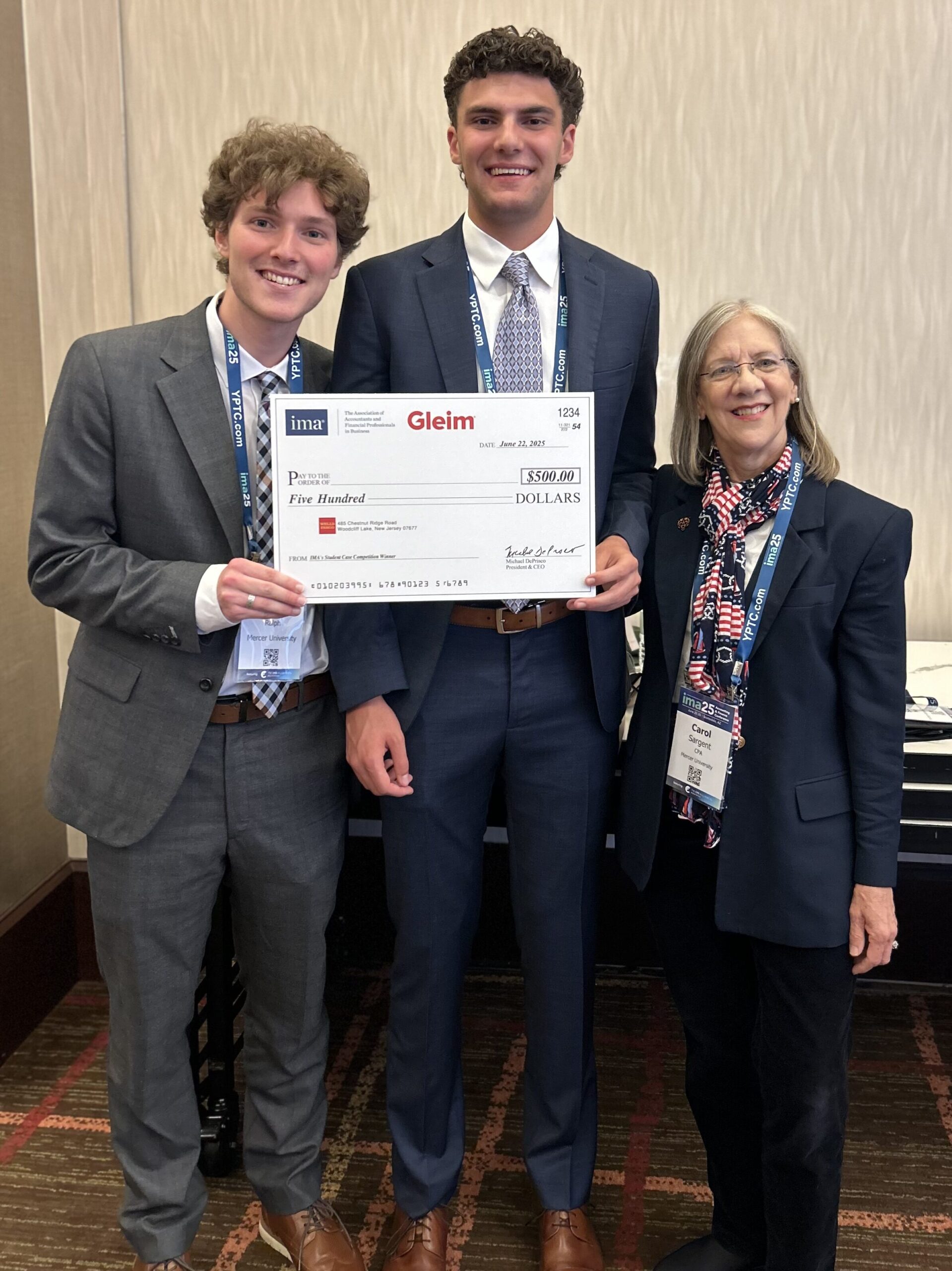 Two young men in suits and an older woman stand indoors, holding a large $500 check from Gleim and Ima.