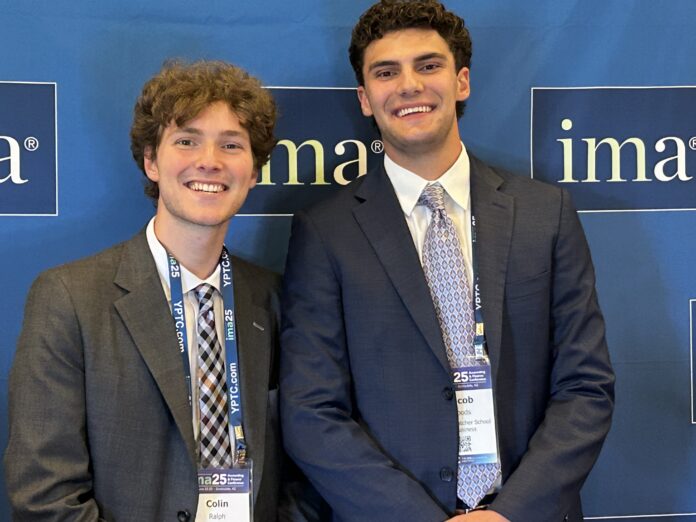 Two young men in suits smiling and standing in front of a blue IMA conference backdrop with name badges visible.