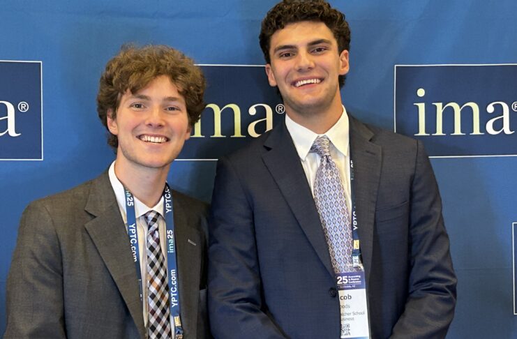Two young men in suits smiling and standing in front of a blue IMA conference backdrop with name badges visible.
