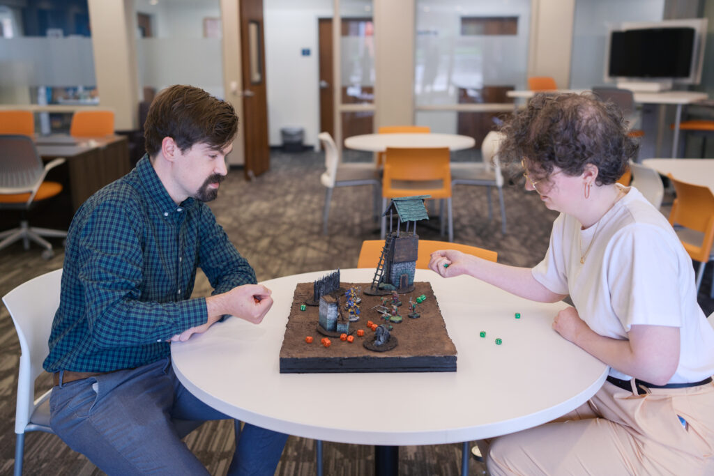 Two people sit at a table playing a tabletop game with miniatures, dice, and terrain props in an office setting.