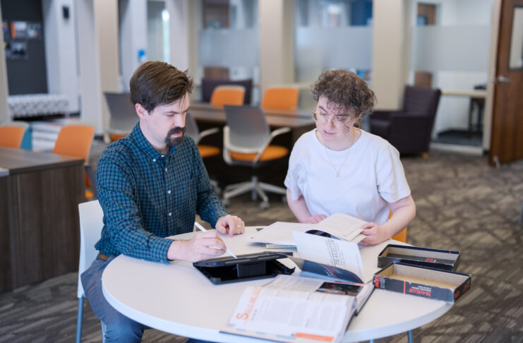 Two people sit at a round table in an office, reading books and taking notes together.