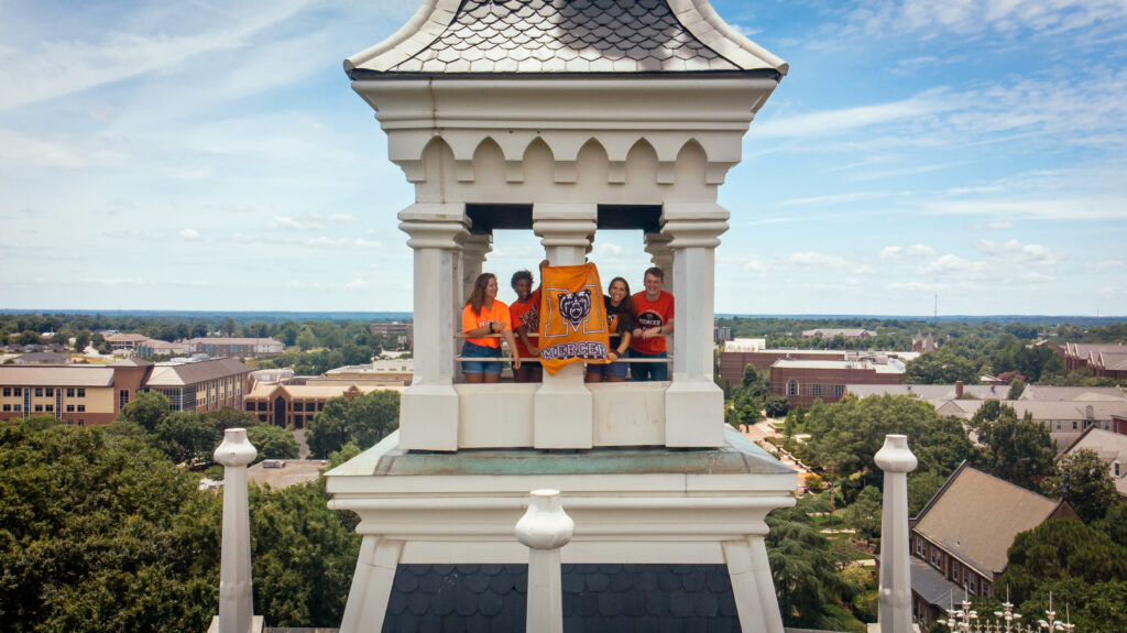 Four people stand in a white clocktower holding a Mercer University flag, with campus buildings visible below.
