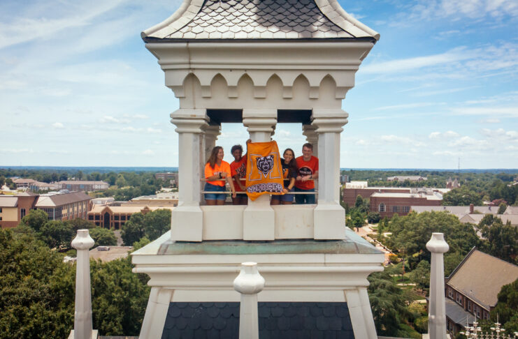 Four people stand in a white clocktower holding a Mercer University flag, with campus buildings visible below.