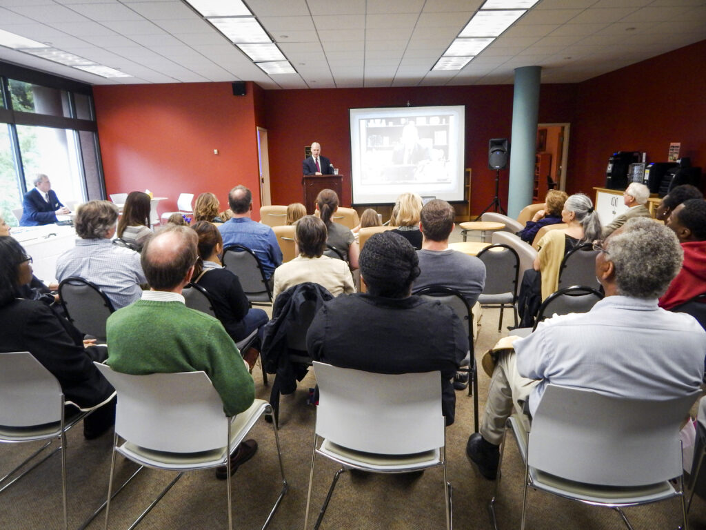 A group of people sit facing a speaker giving a presentation in a conference room with a projector screen.