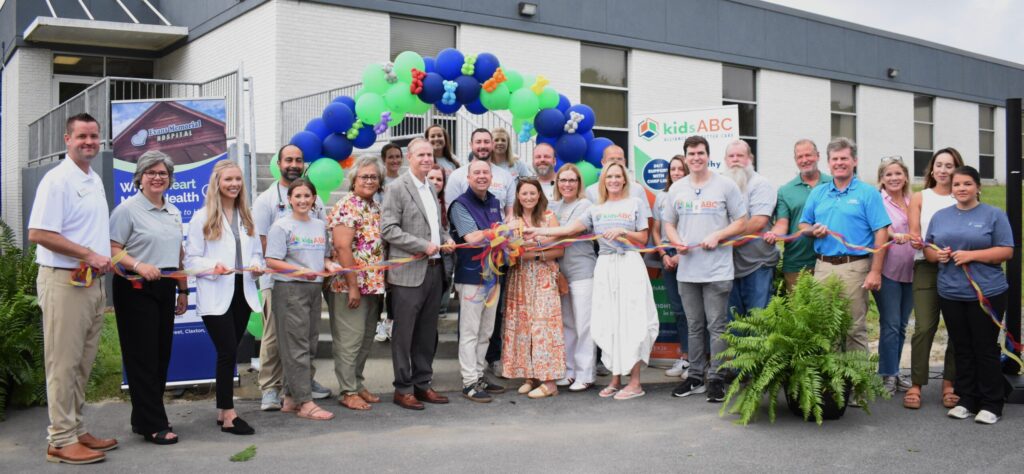 A group of people stand in front of a building holding a ribbon for a ribbon-cutting ceremony with balloons overhead.